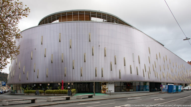 Large modern building with broad curved roof, pale vertical cladding, narrow slit windows, and glazed street-level frontage.