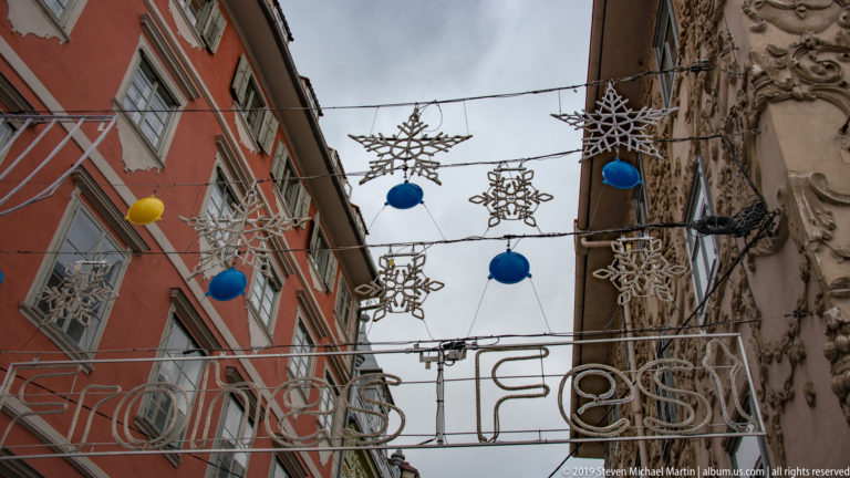 Overhead festive light decorations and colorful spheres strung between historic buildings above a narrow Graz street.