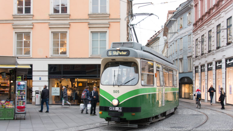 Green-and-cream Graz tram number 505 turning through a shopping street with pedestrians and overhead wires.