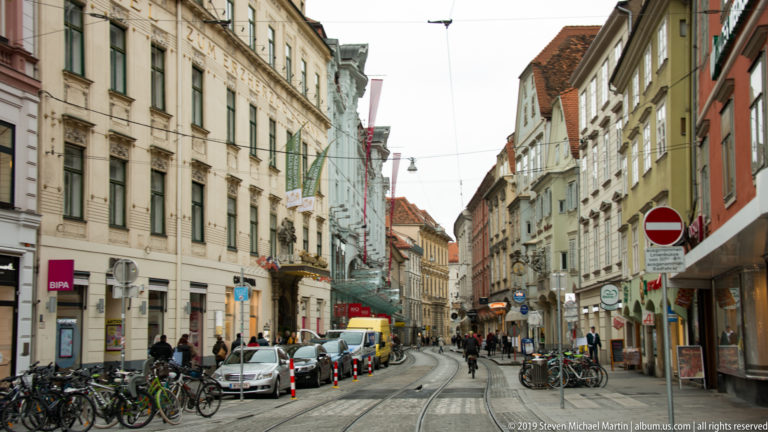 Tram tracks, bicycles and cars along a narrow street lined with historic multi-story buildings in Graz.
