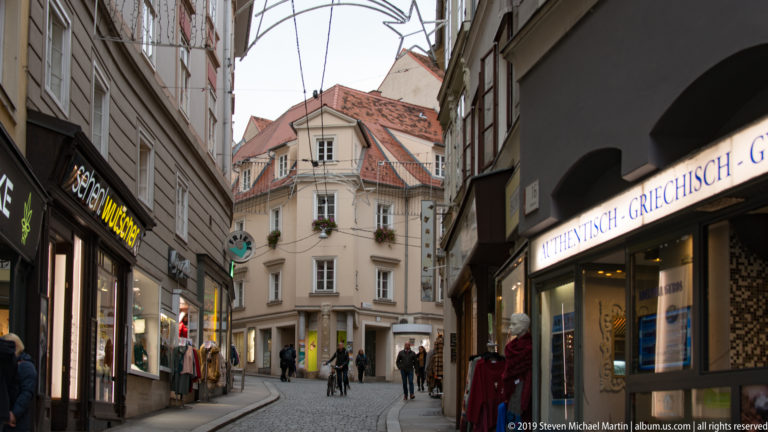 Curving narrow lane with shops, pedestrians and a pale red-roofed corner building in central Graz.