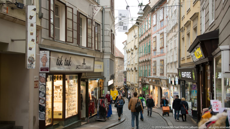 Narrow pedestrian shopping street in Graz with historic façades, storefronts, signs and people walking between the buildings.