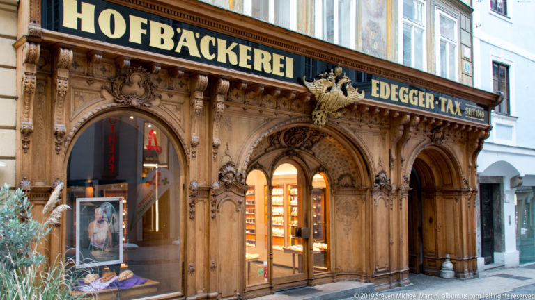 Ornately carved dark wooden Edegger-Tax bakery façade with arched windows, columns and heraldic decoration.