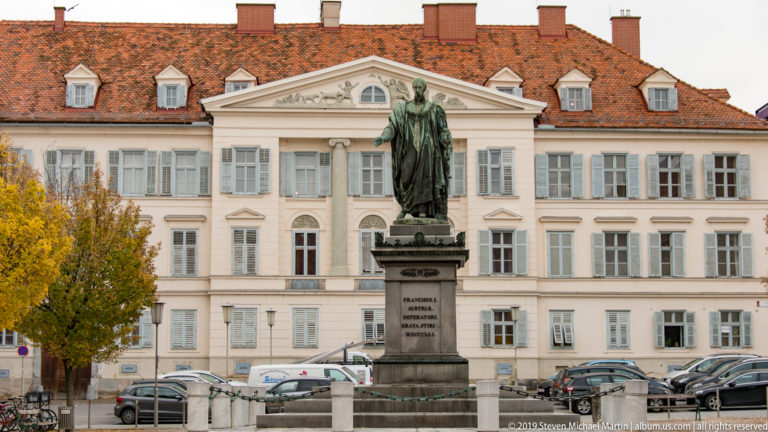 Bronze robed figure on a tall stone pedestal in front of a pale classical building in Graz.