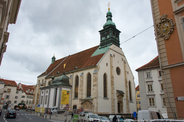 Large historic church with pointed windows, red tile roof and green copper tower at a street corner in Graz.