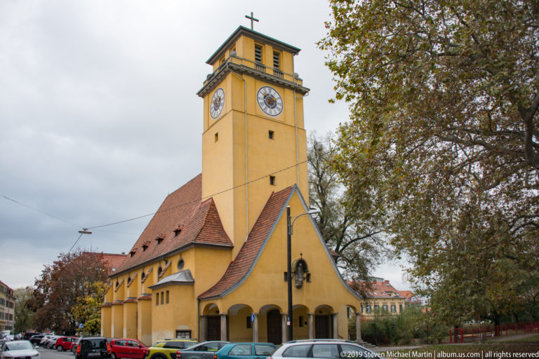 Yellow church with long tiled nave, square clock tower, clock faces, cross, arched porch, trees, and parked cars.