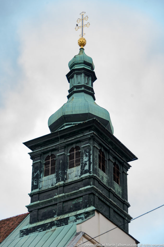 Dark green copper tower with open belfry, tiered roof and gilded finial against a pale gray sky.