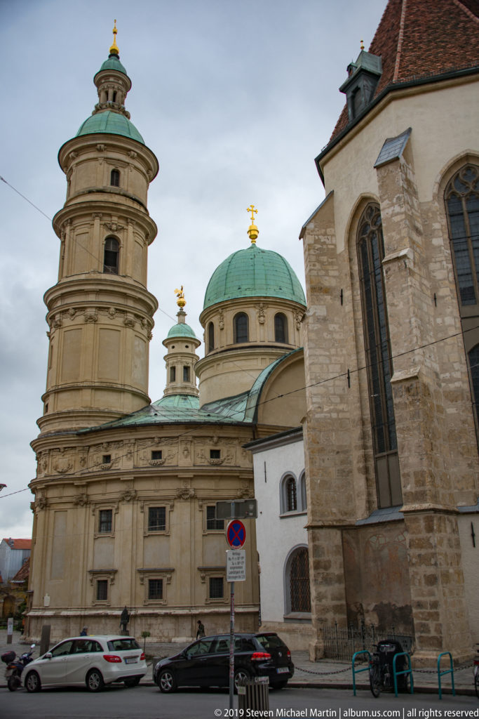 Stone church buildings with green copper domes and lanterns beside the Gothic wall of Graz Cathedral under an overcast sky.