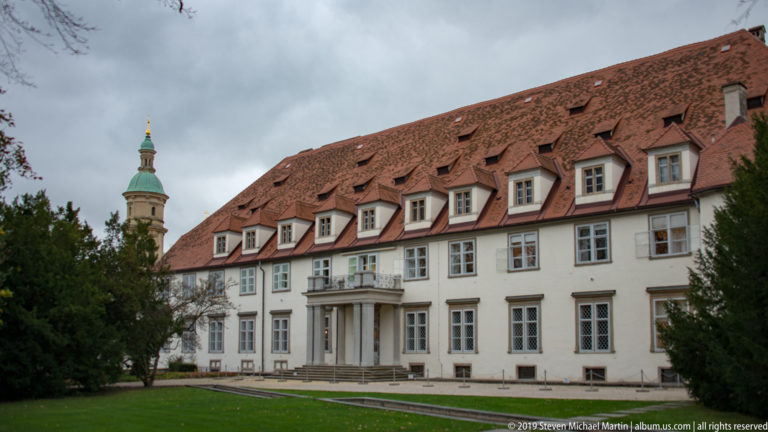 Long white historic building with red dormered roof, central porch, lawn, and landscaped foreground.