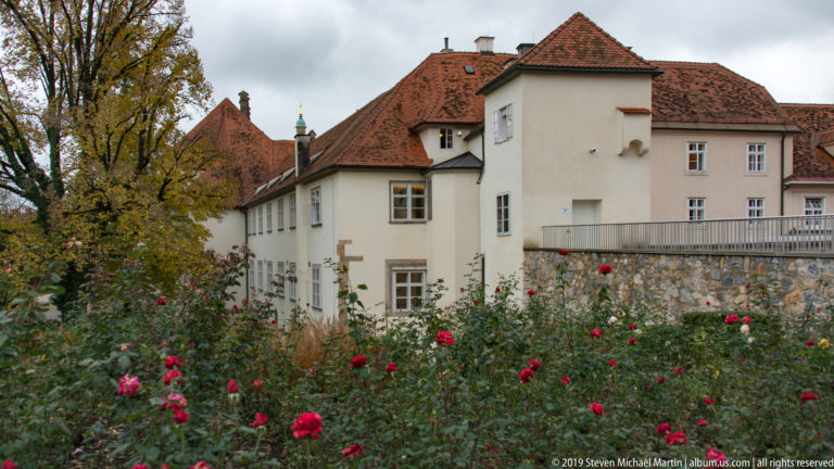 White historic building with red tile roofs behind a rose garden and stone wall in Graz.