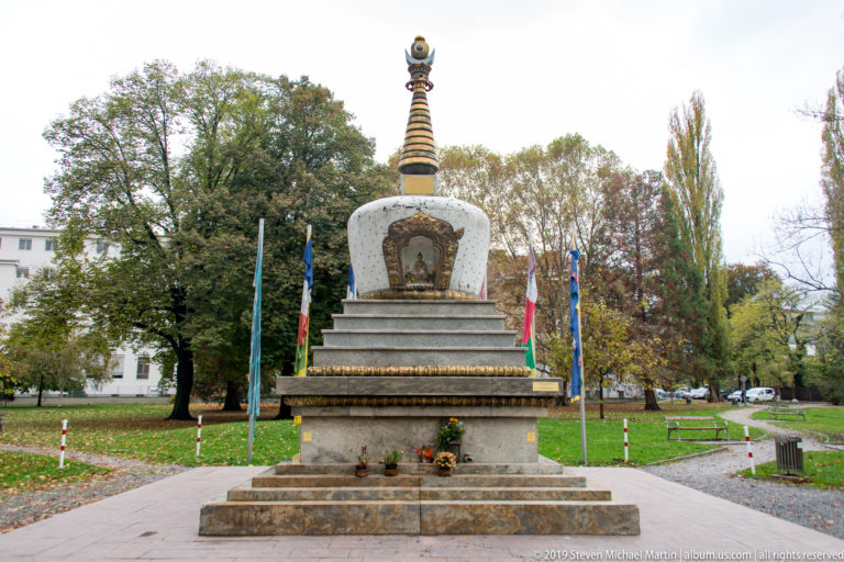 White and gold Tibetan-style Buddhist stupa on a stepped stone base with prayer flags and trees behind.