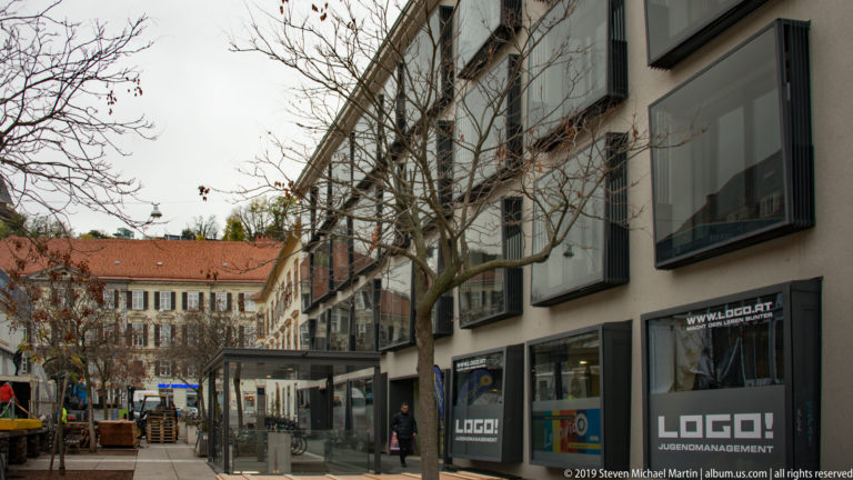 Modern glass-fronted commercial building beside trees and café seating on a Graz pedestrian street.