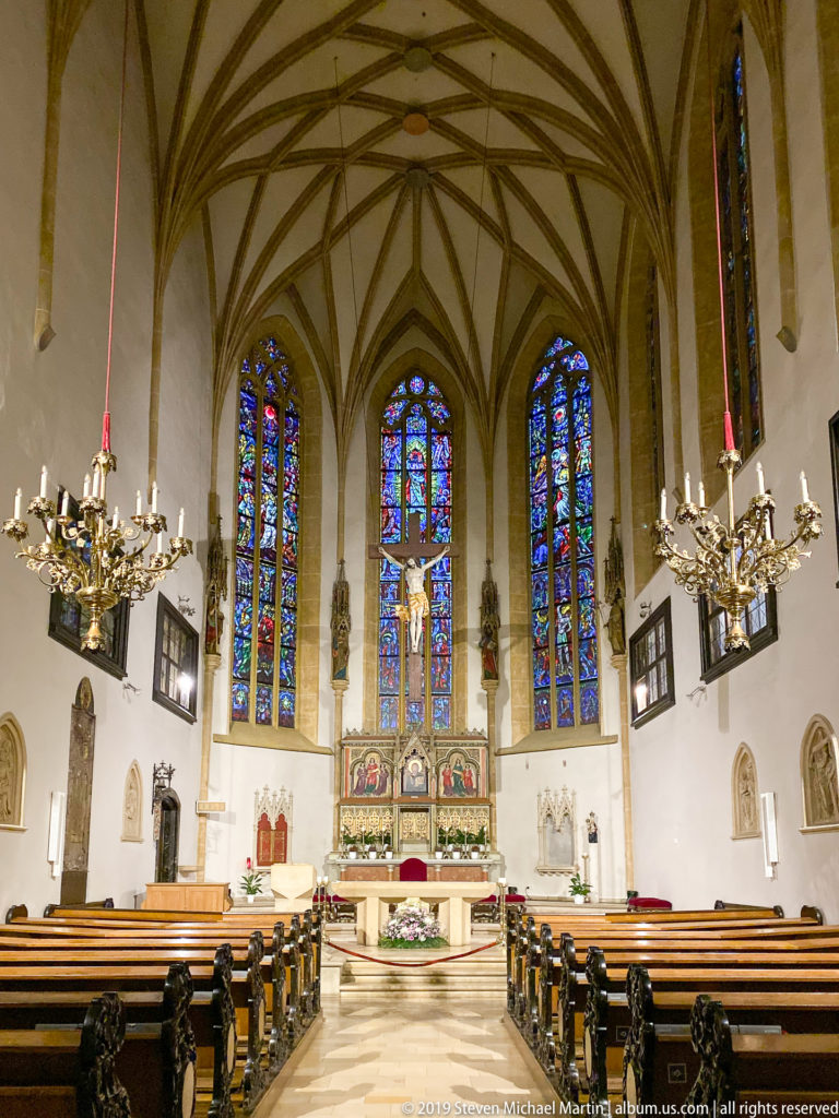 Symmetrical church sanctuary with crucifix, altar, stained-glass windows, chandeliers, and pews.