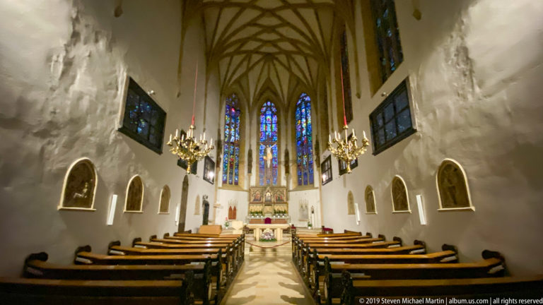 Wide church nave with wooden pews, chandeliers, ribbed ceiling, altar, and tall stained-glass windows.