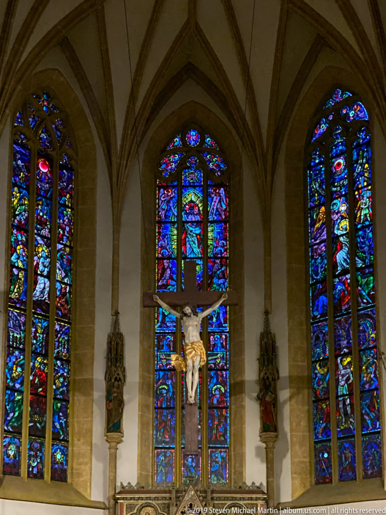 Frontal church interior with crucifix, vivid stained glass, and carved saints between pointed windows.