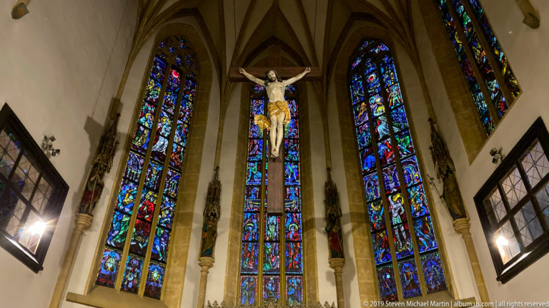 Church sanctuary with ribbed vaults, central crucifix, statues, and three tall stained-glass windows.