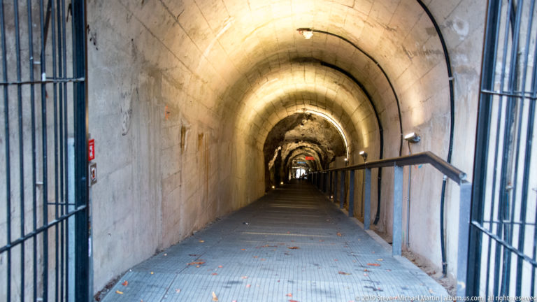 Long illuminated Schlossberg tunnel with concrete vault, exposed rock, railings, grilles, and rising walkway.