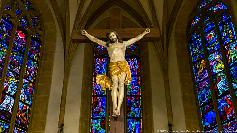 Crucifix with figure of Christ centered before tall blue, red, and violet stained-glass windows.
