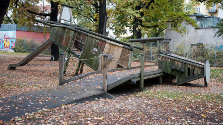 Weathered wooden playground structure with sloped ramps, rails, and slide among fallen leaves and trees.