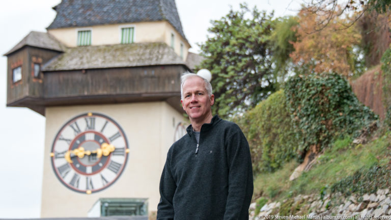 Man in dark sweater standing in front of the blurred Graz Uhrturm and autumn hillside vegetation.