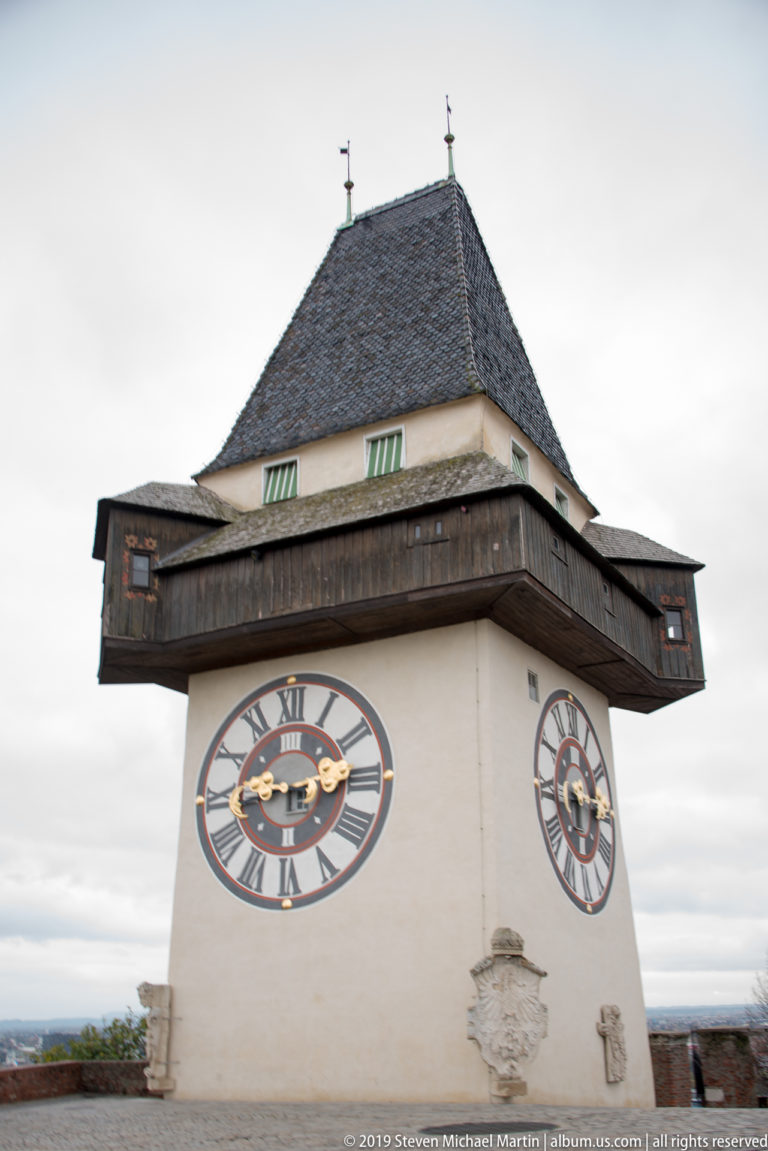 Three-quarter view of the Graz Uhrturm with two clock faces, timber gallery, and steep shingled roof.
