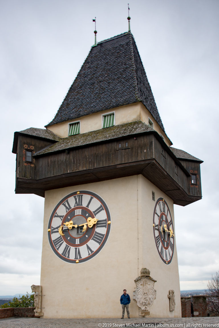 Oblique view of the Graz Uhrturm showing two clock faces, dark timber gallery, steep roof, and visitor.