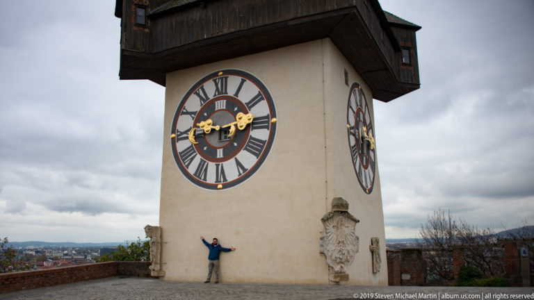 Graz Uhrturm with large clock face, broad wooden gallery, steep roof, and a visitor at its base.