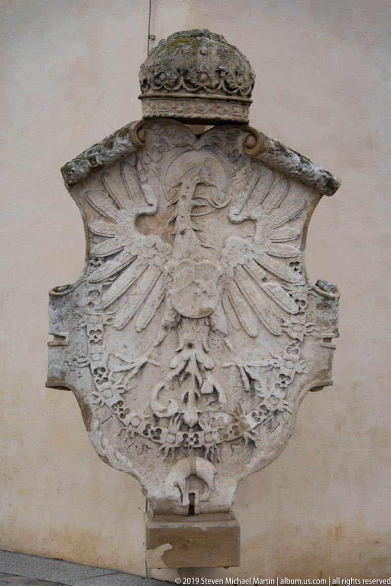 Weathered stone relief of a crowned double-headed eagle with shield mounted on a pale wall.