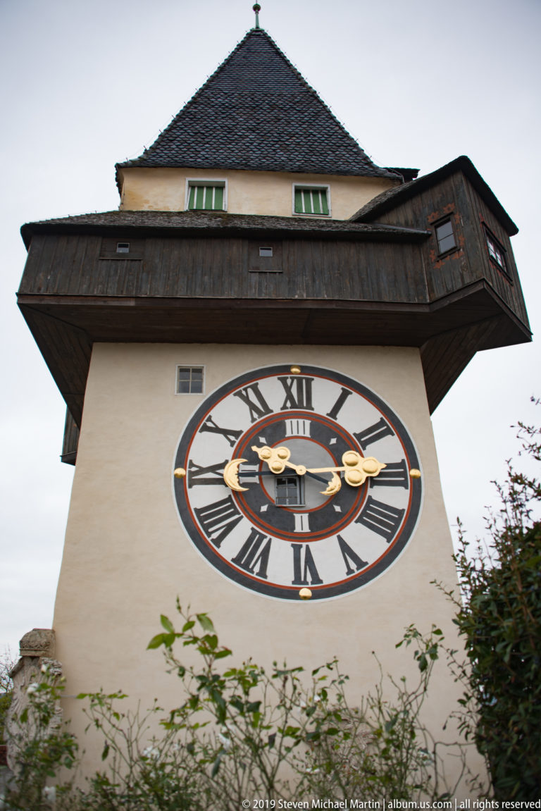 Large Roman-numeral clock face beneath deep wooden eaves on the Graz Uhrturm.