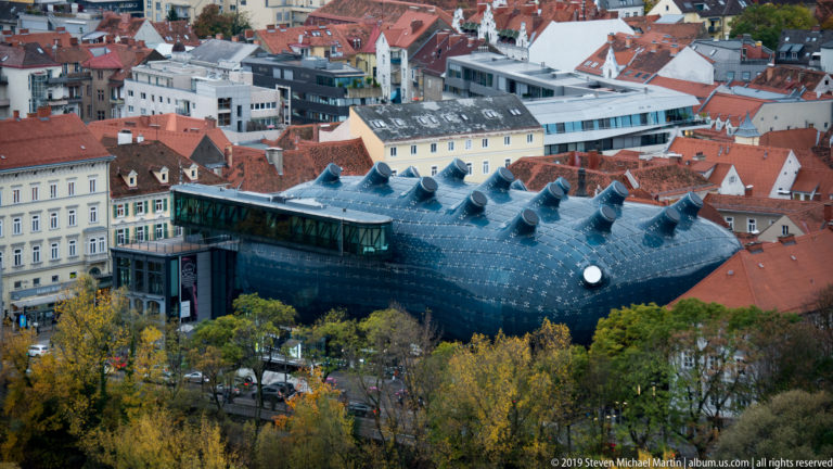 Kunsthaus Graz with dark curved exterior and projecting skylights amid red-roofed historic buildings.