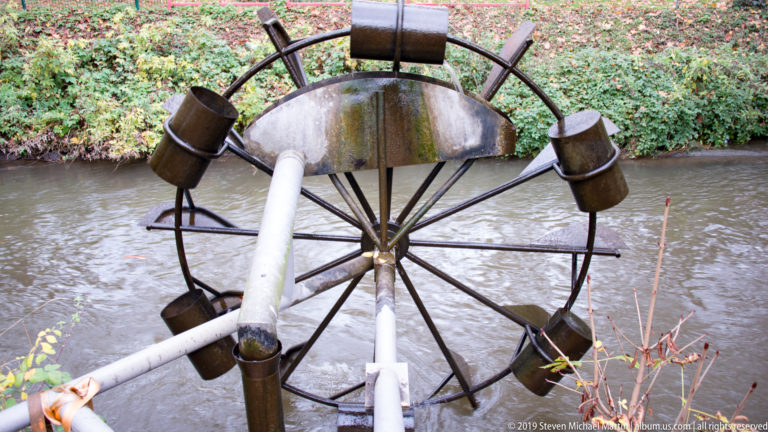 Metal bucket water wheel beside a narrow stream, with a pipe aimed toward the wheel and vegetation behind.
