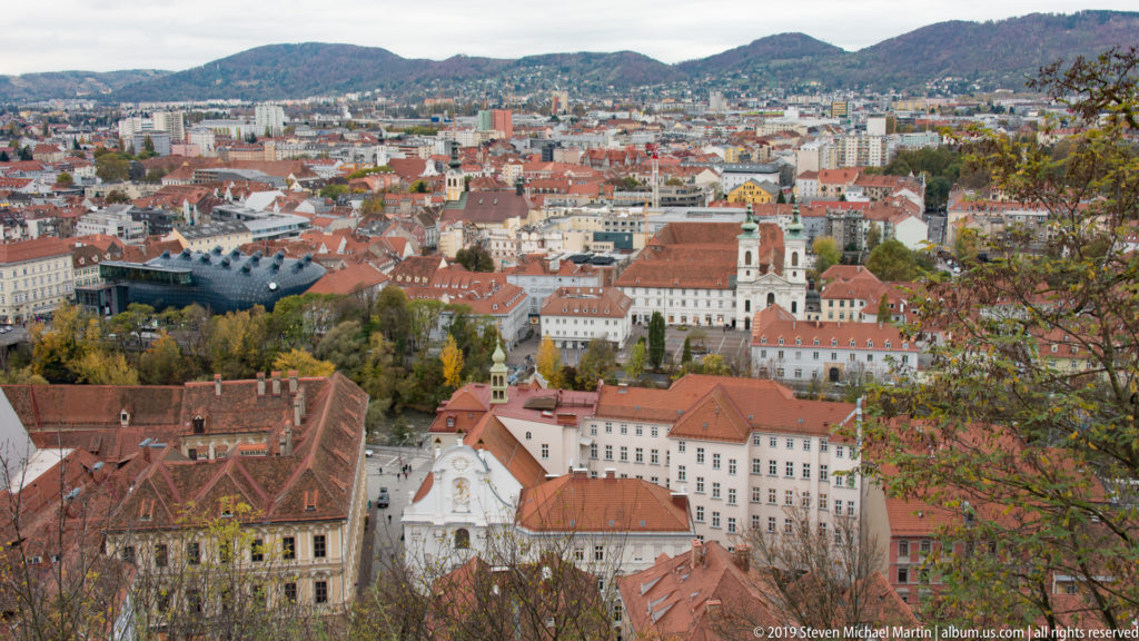 Elevated panorama of Graz with red roofs, church towers, dense urban blocks, and wooded hills beyond.
