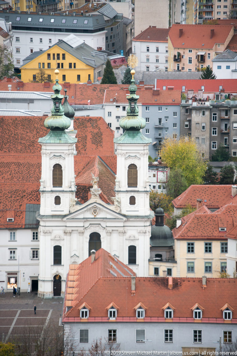 White twin-towered Baroque church with green copper caps amid red-tiled roofs in central Graz.