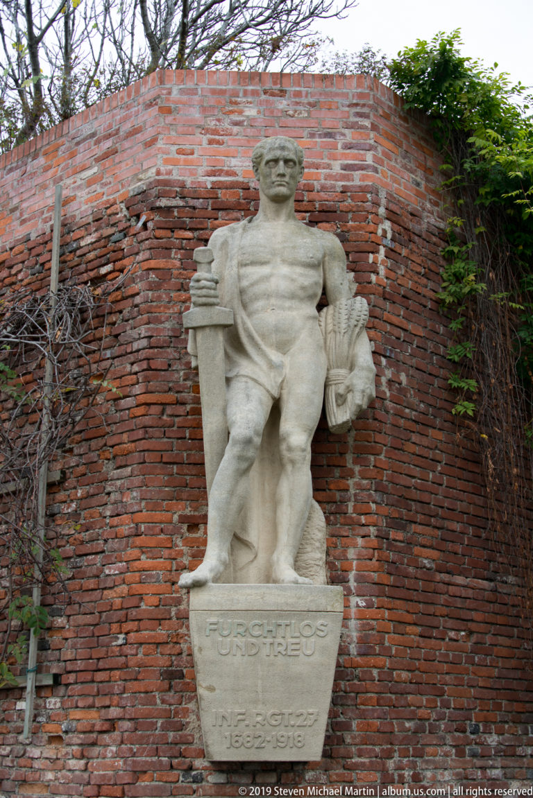 Stone statue of Hercules with club and drapery on an inscribed pedestal, set against a curved red-brick wall on Schlossberg.