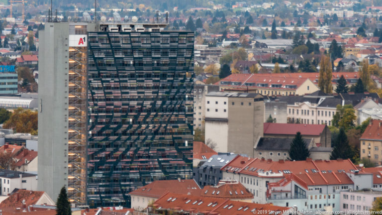 Telephoto view from Schlossberg toward the dark biomorphic Kunsthaus Graz amid red-roofed historic buildings and modern city blocks.