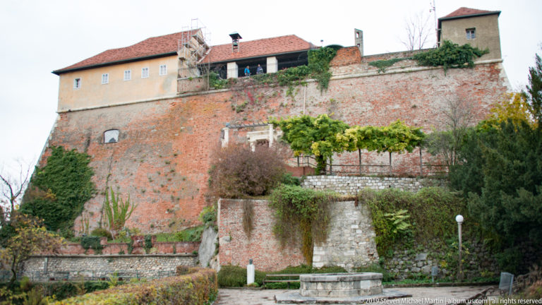 Tall red-brick Schlossberg walls above terraced gardens and stone retaining walls, with pale buildings and a red-tiled roof on top.