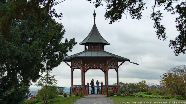 Open wooden pavilion with steep roof and decorative railings on Schlossberg, with visitors standing at the overlook.