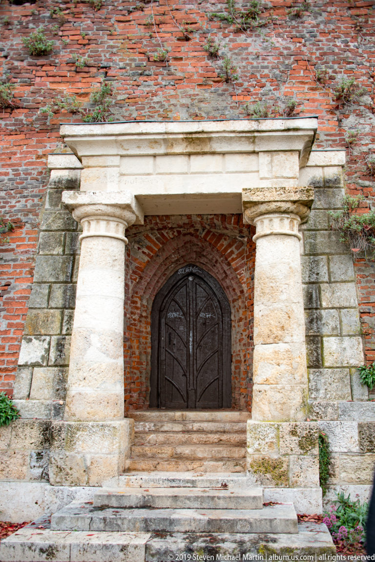 Stone steps leading to an arched dark wooden door set in a red-brick Schlossberg wall between massive pale stone piers.