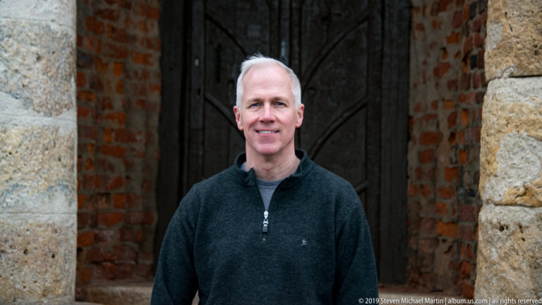 Man in a dark sweater standing before a wooden doorway framed by rough stone and exposed red brick on Schlossberg.