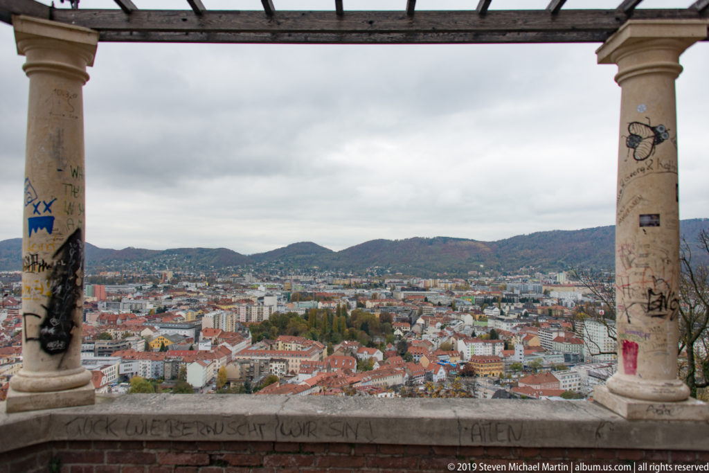 Panoramic view over Graz from Schlossberg, framed by columns and a beam, with red roofs and wooded hills beyond.