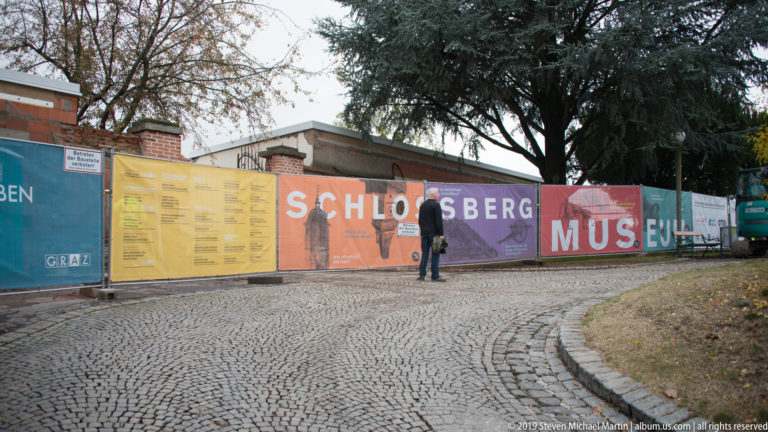 Colorful barrier reading SCHLOSSBERG MUSEUM along a cobbled path, with a pedestrian and trees behind it.