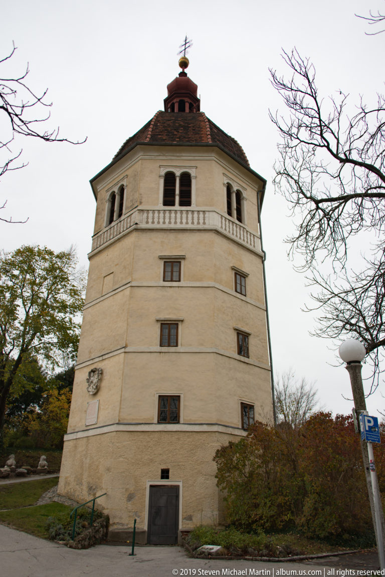 Pale octagonal Glockenturm on Graz Schlossberg with small windows, open belfry arches, polygonal roof, and weather vane.