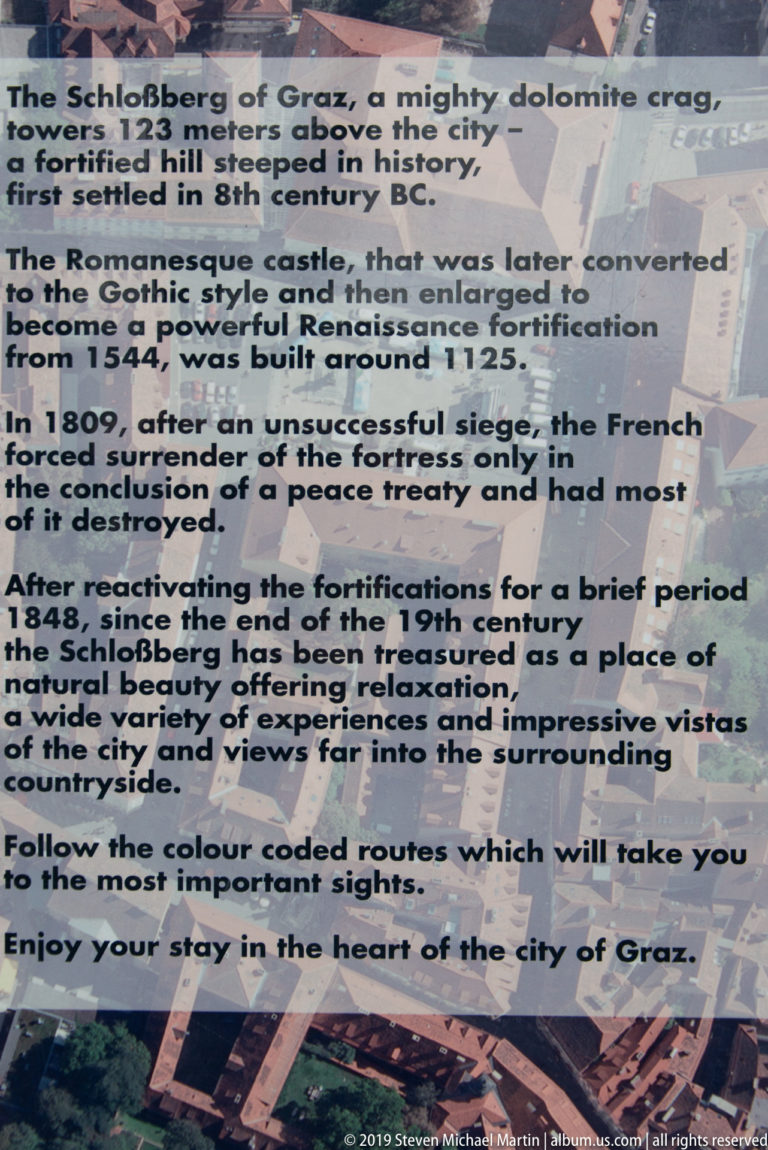 English interpretive panel on Schlossberg describing its castle, fortress, 1809 siege, demolition, park, and visitor routes.
