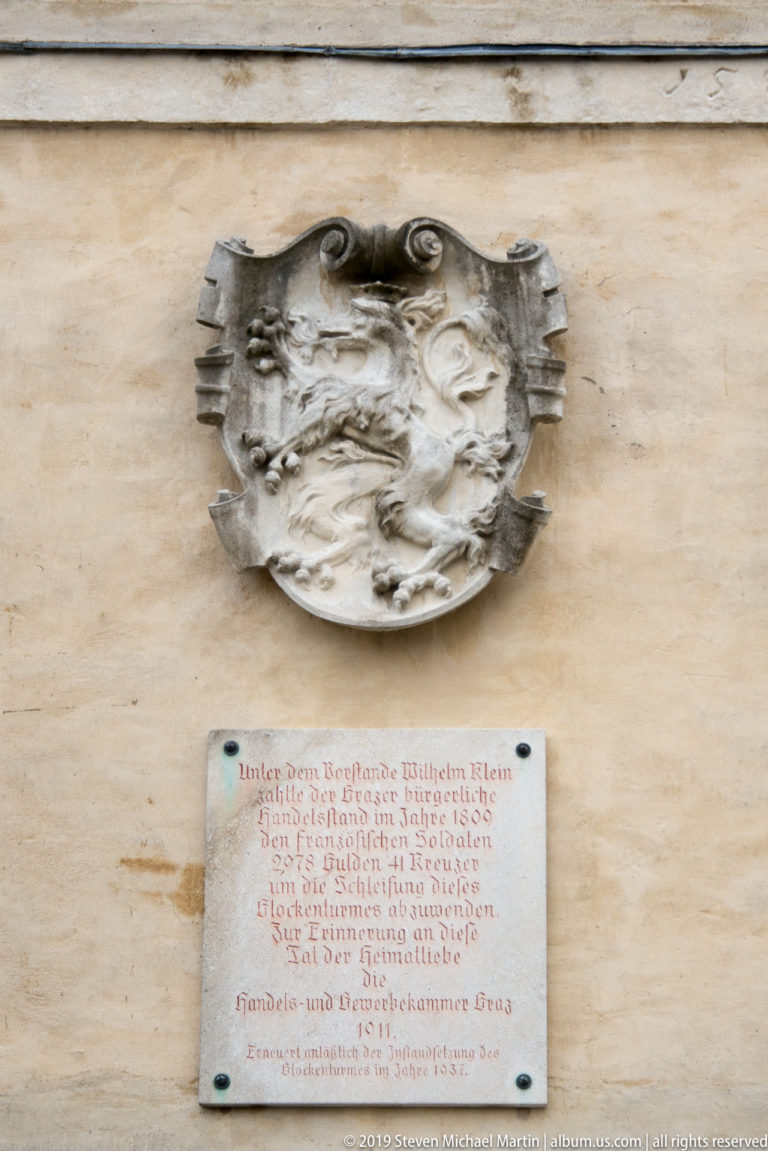 Weathered figurative relief above a rectangular German inscription plaque on a pale wall at Graz Schlossberg.