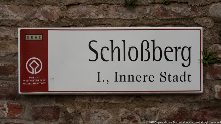White sign reading Schloßberg, I., Innere Stadt with UNESCO World Heritage emblem, mounted on a rough brick-and-stone wall.
