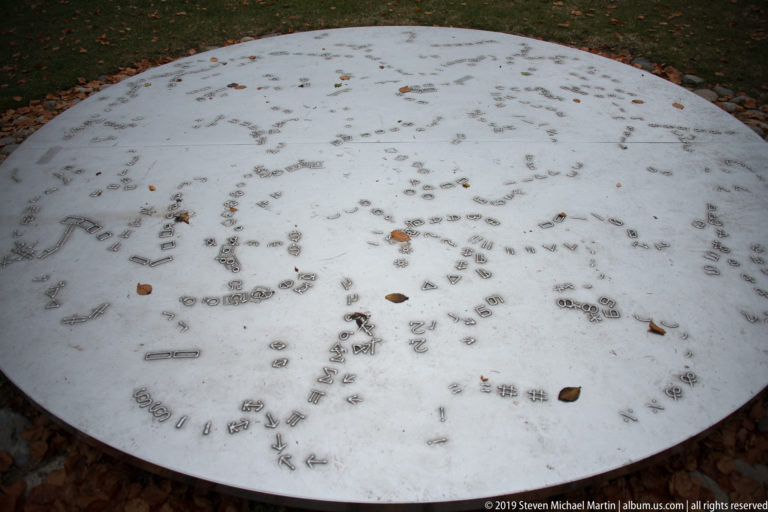 Large circular pale metal disk outdoors on Schlossberg, densely engraved with names and small marks and scattered with fallen leaves.