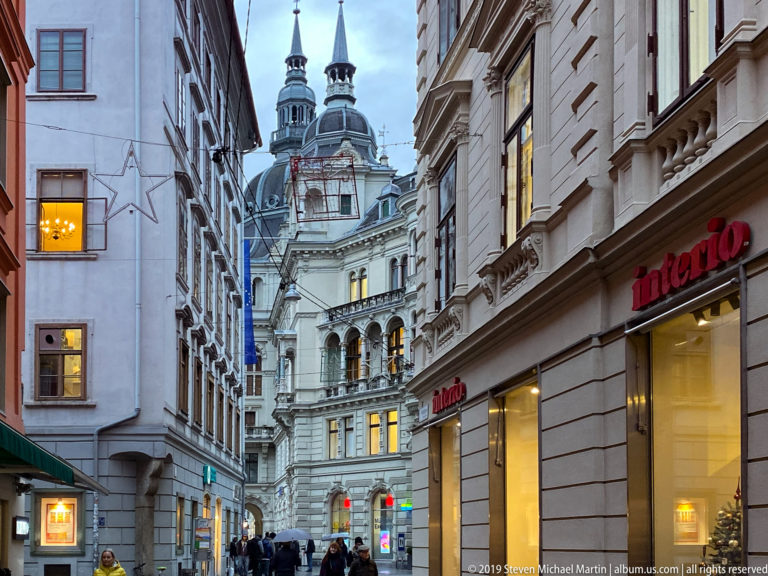 Narrow Graz street with pedestrians and lit shops leading toward an ornate church façade with twin slender towers.