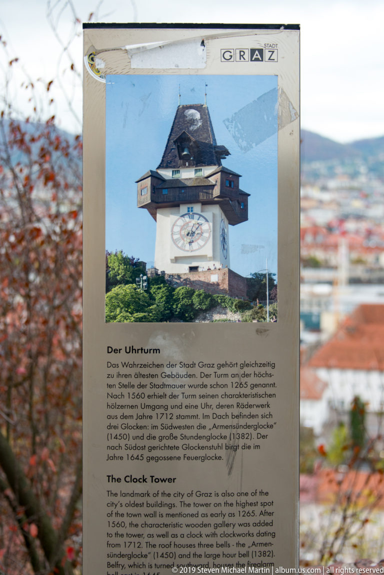 Bilingual Der Uhrturm / The Clock Tower interpretive sign with a tower photograph, set against a blurred view of Graz.