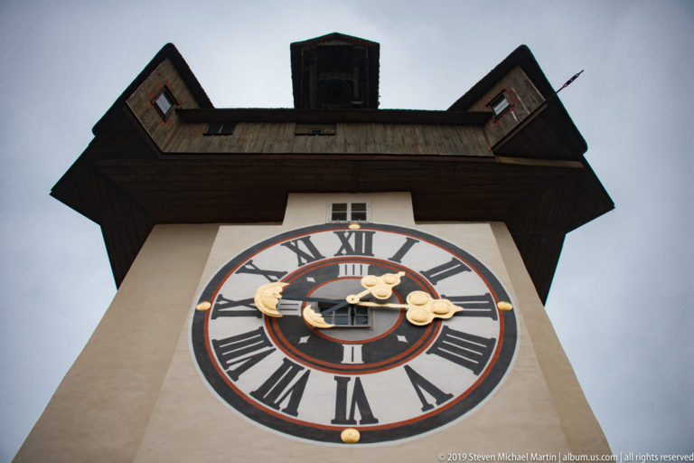 Low-angle close view of the Grazer Uhrturm clock face with Roman numerals, gilded hands, painted rings, and projecting roof.