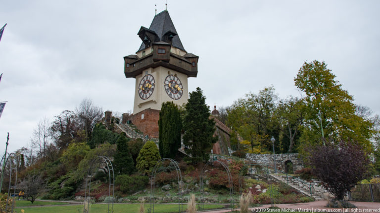 Grazer Uhrturm on Schlossberg with large clock faces, broad wooden eaves, steep roof, autumn trees, and landscaped paths.